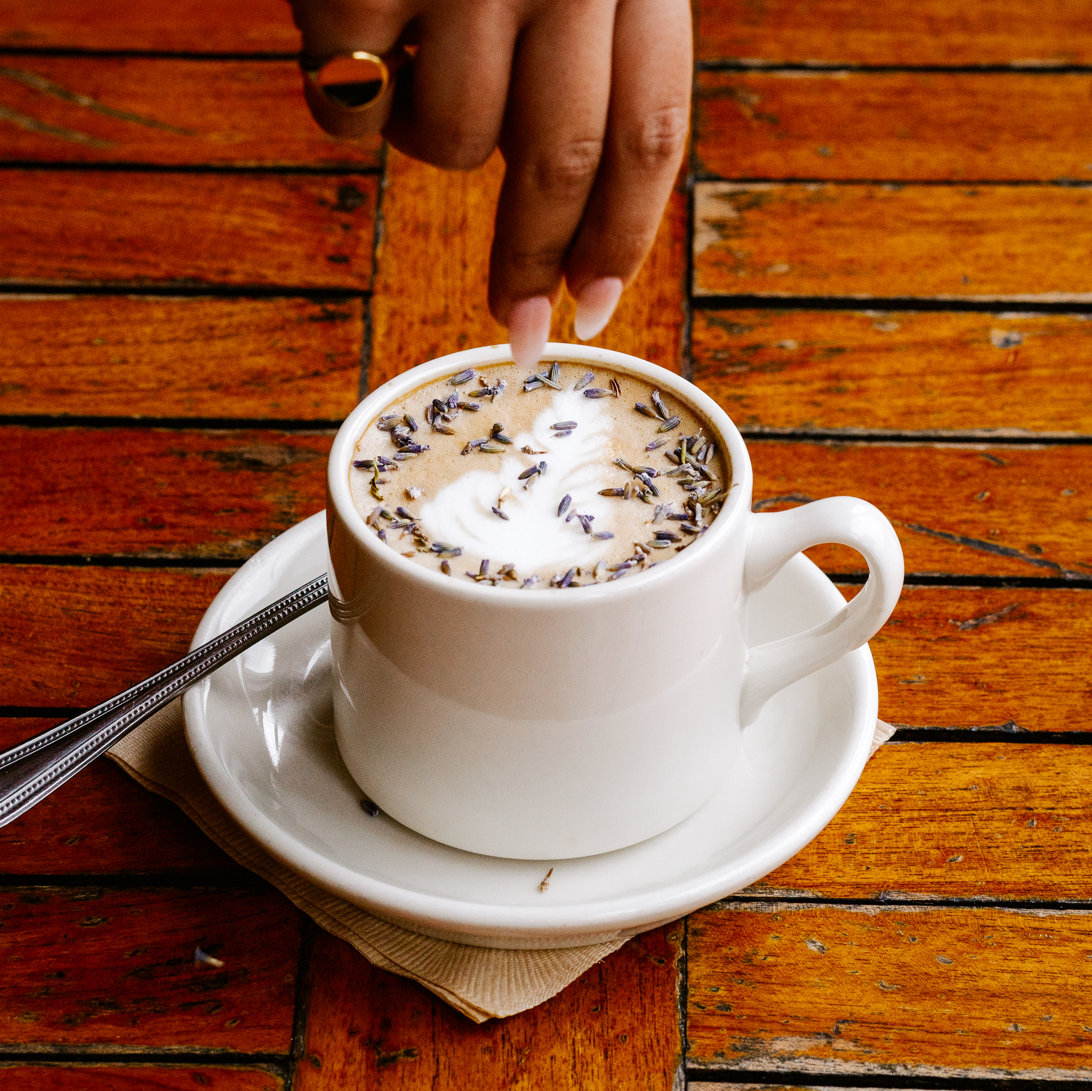 A well-manicured hand drops lavender leaves onto a freshly brewed latte in a mug.
