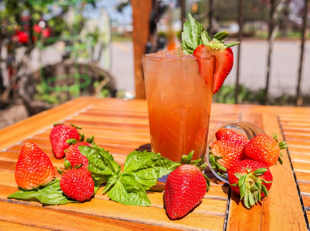 A bright pink cocktail with a strawberry garnish, surrounded by fresh strawberries and basil.