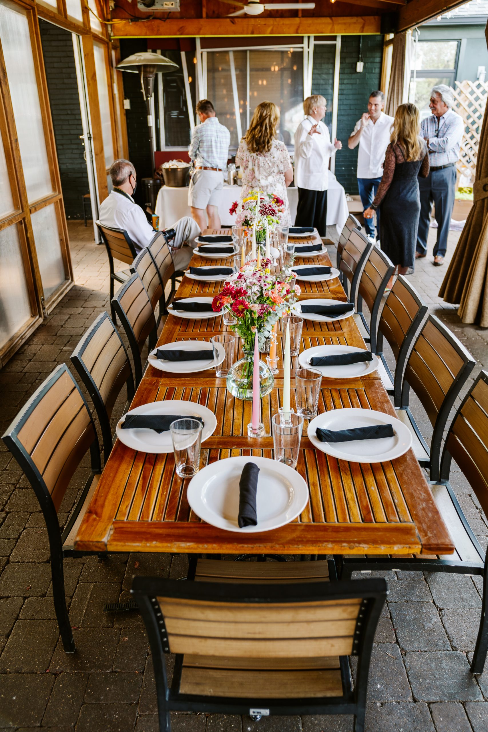A long table set up in an outer patio with people standing and mingling.