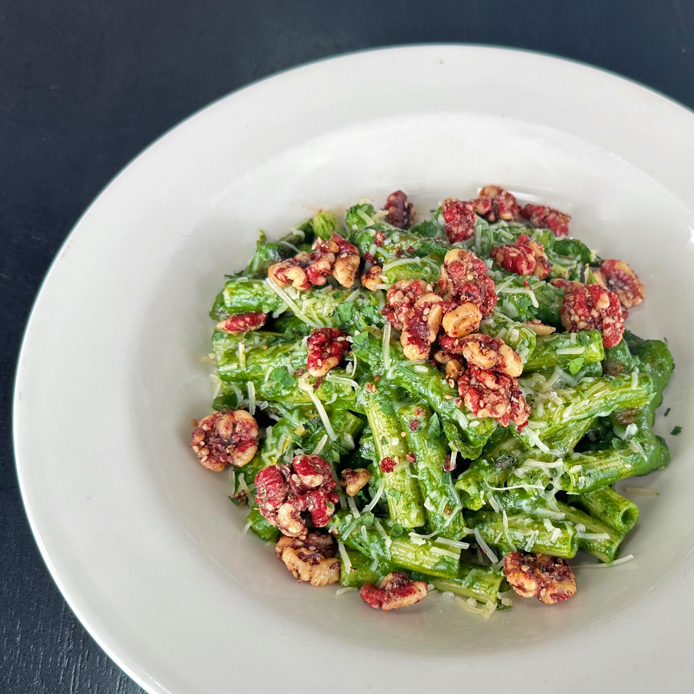 A bowl full of pasta tossed in a bright green pesto, topped with shredded parmesan and toasted red walnuts.