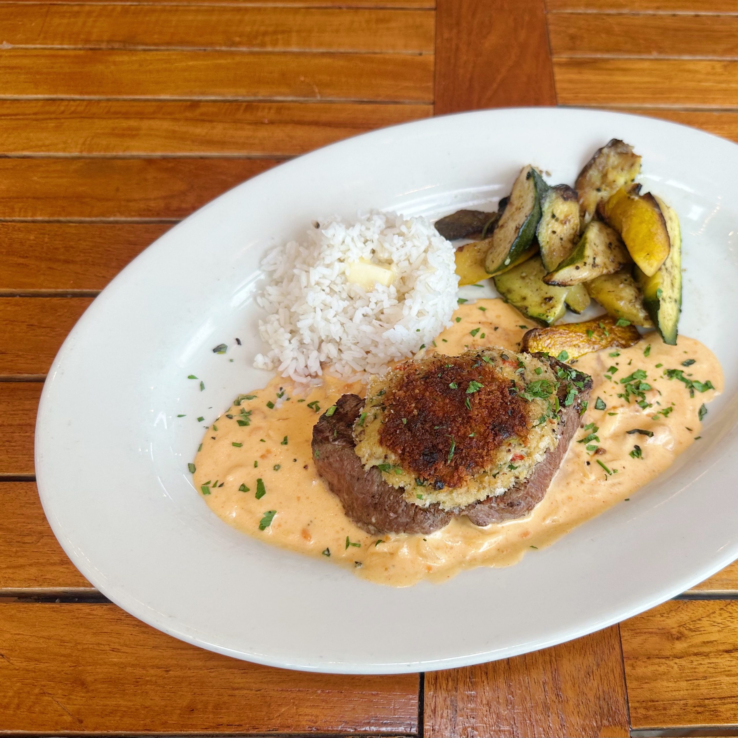 A steak topped with a crab cake, plated with a creamy sauce, side of rice, and grilled zucchini.