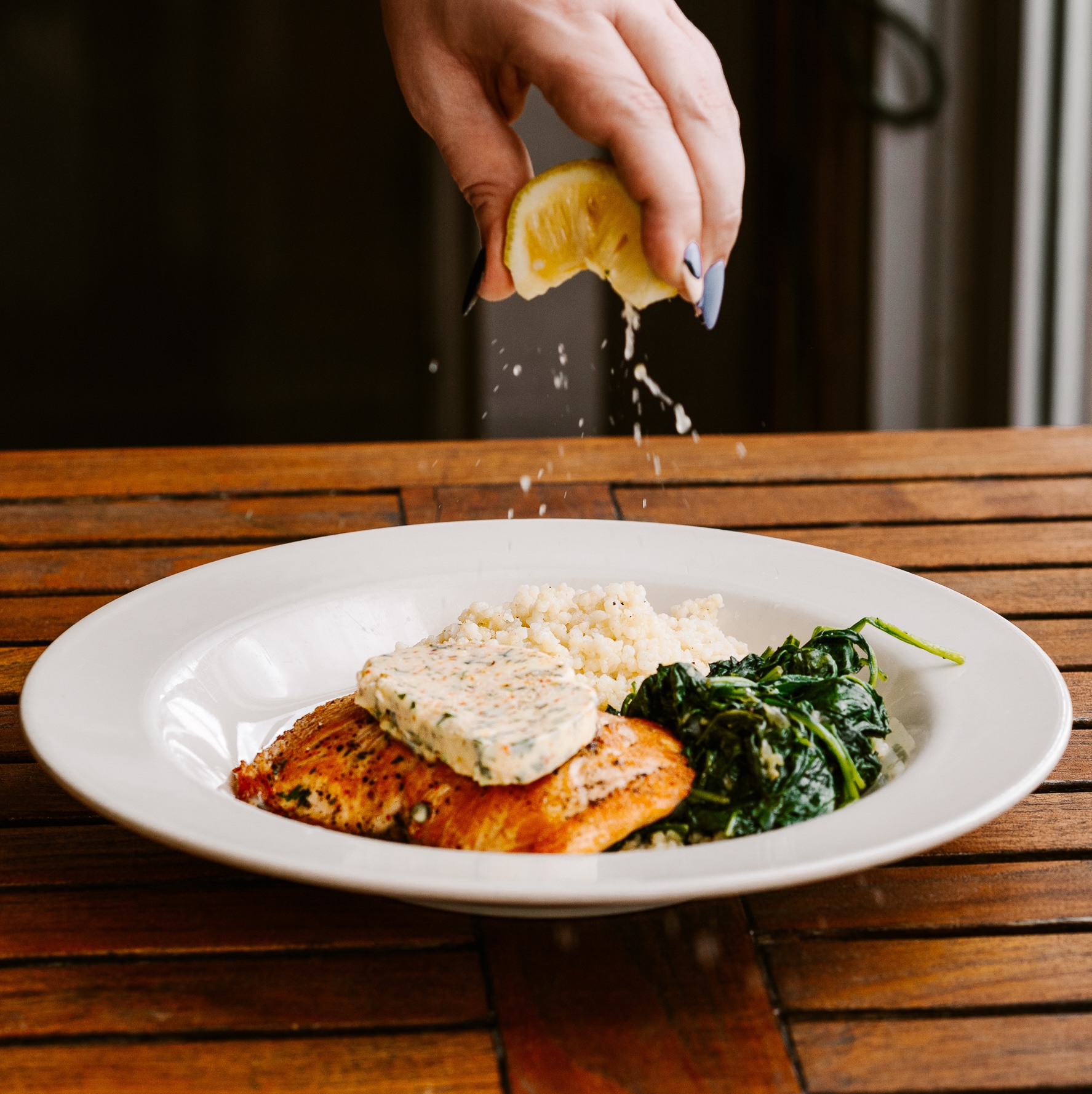 A grilled salon dish with herbed butter on top, with a hand squeezing lemon over the fish.